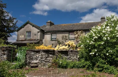 An exterior view of Hill Top, a traditional grey stone farmhouse with green-trimmed windows under a blue sky. A low dry-stone wall and a bright green metal gate stand in the foreground, surrounded by lush green bushes and blooming white and yellow flowers.