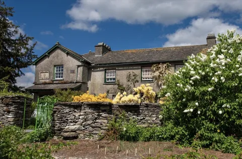 An exterior view of Hill Top, a traditional grey stone farmhouse with green-trimmed windows under a blue sky. A low dry-stone wall and a bright green metal gate stand in the foreground, surrounded by lush green bushes and blooming white and yellow flowers.