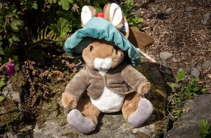 A plush soft toy of Benjamin Bunny sitting on a mossy rock in a garden. The rabbit wears his signature oversized teal tam-o'-shanter hat with a red pom-pom and a brown jacket. Green garden plants and a single pink flower are visible in the background.