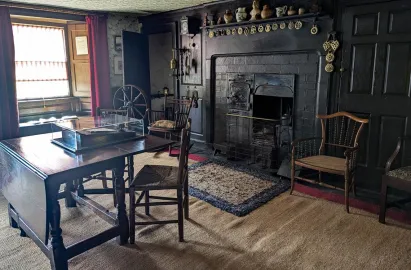The interior Entrance Hall of Hill Top, featuring a dark wood-paneled wall with a large black cast-iron range fireplace. A wooden gate-leg table stands in the foreground, and various antique chairs, a spinning wheel, and horse brasses decorate the room. Sunlight streams in from a window with deep red curtains on the left.