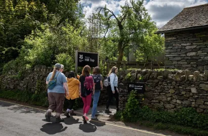 A group of visitors with backpacks walks along a paved road toward the stone-walled entrance of Hill Top. A prominent black National Trust sign reads "Welcome to Hill Top." A second smaller sign on the dry-stone wall points toward the "Car park and information" area under a canopy of lush green trees and a bright, cloudy sky.