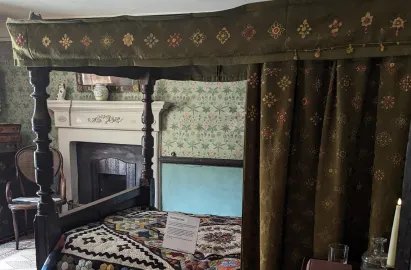 An interior view of a bedroom at Hill Top featuring a dark wood four-poster bed. The bed is dressed with a heavy green canopy and curtains embroidered with floral patterns, and a colorful patchwork "Wedding Quilt." In the background, a white mantelpiece sits against green floral wallpaper, and a lit candle glows in the foreground.