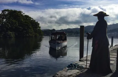 A silhouette of a re-enactment of Beatrix Potter, dressed in a long coat and wide-brimmed hat with a walking stick, stands on a stone lakeside jetty. In the center of the frame, a vintage-style white motor launch boat approaches the shore across the calm water of Lake Windermere under a cloudy sky.