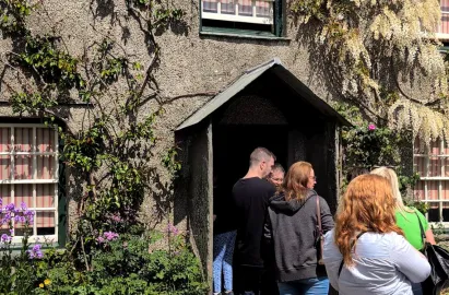 A close-up view of a group of visitors gathered at the entrance of Hill Top. They are standing in front of the small, slate-roofed porch, waiting to enter the dark doorway of the grey stone farmhouse. To the left, purple wildflowers and climbing green vines decorate the wall next to a white-framed window with red-striped curtains.