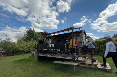 A rustic outdoor café kiosk named "Joey’s Lake District" situated on a grassy hill. The wooden kiosk features a black banner advertising "Bar, Coffee, and Food." Several visitors are lined up at the counter or walking up the wooden pallet steps under a bright blue sky with fluffy white clouds.