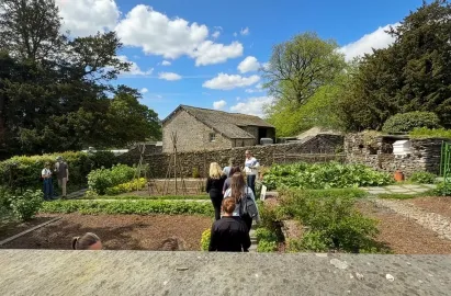 A high-angle view looking over a stone wall into the Hill Top vegetable garden. Several visitors walk along a slate path between raised planting beds and tall wooden bean poles. In the background, a large traditional stone barn stands under a bright blue sky with scattered white clouds, surrounded by mature green trees.