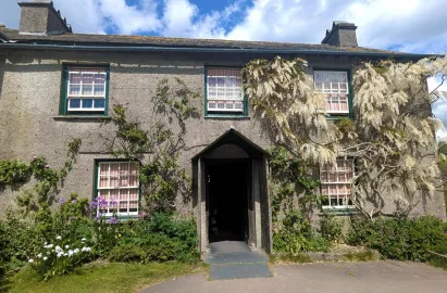 A straight-on exterior view of the front of Hill Top farmhouse. The grey stone building features a central gabled porch with an open dark doorway. Climbing plants with white flowers and purple wildflowers frame the green-trimmed sash windows, which are draped with red and white striped curtains.