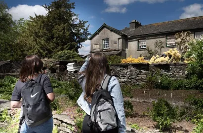 Two visitors with backpacks stand in a lush garden, viewed from behind, looking toward the Hill Top farmhouse. The historic stone building is framed by a tall evergreen tree, blooming yellow flowers, and a traditional dry-stone wall under a bright, partly cloudy sky.