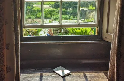 An interior view looking out through a white-framed sash window of the farmhouse. On the dark, wooden deep-set windowsill lies a small copy of "The Tale of Two Bad Mice." Through the glass, a sunny garden with stone paths, lush green ferns, and visitors walking by is visible. Floral patterned curtains frame the shot on both sides.