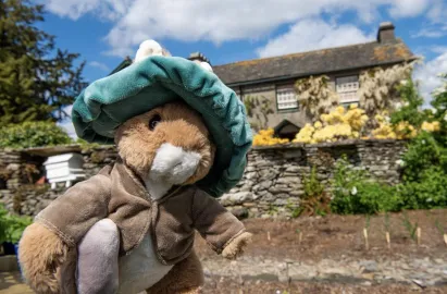 A close-up, slightly low-angle shot of a Benjamin Bunny plush toy wearing a teal hat and brown jacket. In the blurred background, the Hill Top farmhouse stands behind a traditional dry-stone wall. The scene is set in a sunny garden under a bright blue sky with scattered clouds.