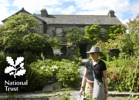 A photo of a lady outside Hill Top Farm with a national Trust logo. Image courtesy of Punktoad on Creative Commons Flickr