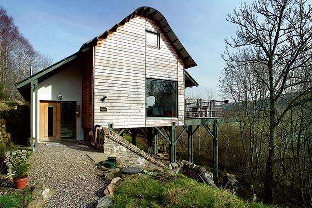 End elevation and front door to a timber framed cladded house with a curved roof on stilts in a Perthshire forest