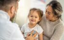 A young girl smiling at the camera and sitting on her mother's lap with a male doctor listening to her lungs with a stethoscope to illustrate paediatricians calling for the roll out of clean air clinics to protect child health