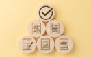 Wooden discs arranged in a pyramid with icons representing checklists, documents, certificates, charts, and a large tick in a circle at the top on a yellow background to illustrate an audit about the use of high-cost therapies such as dupilumab and JAK inhibitors for atopic dermatitis and adherence to NICE criteria for prescribing