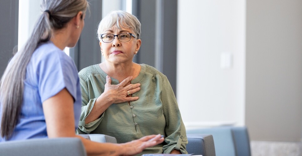An older lady in a green top with her hand over her chest and arthritis in her fingers sitting with a healthcare professional in blue scrubs holding papers and a pen to illustrate pharmacist consultations being found to enhance biologic DMARD therapy