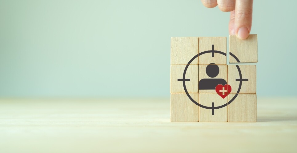 A stack of nine wooden blocks in a square with a hand putting the top right block into place and a silhouette of a person in the middle with a red heart shape overlapped with a white medical cross in the centre and a target shape around them to illustrate a new frontier in precision medicine for complex disease