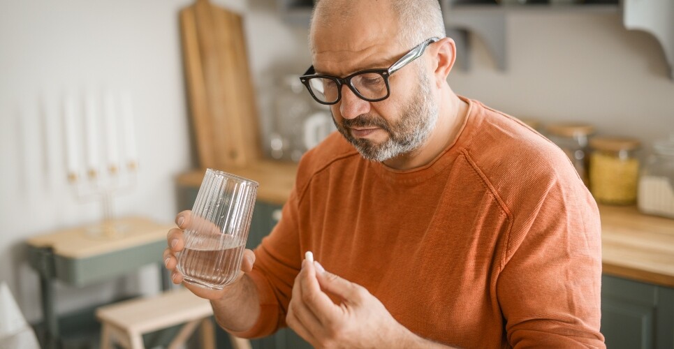 A man in an orange tshirt standing in his kitchen with a glass of water in one hand and a small white pill in the other to illustrate sertraline having an earlier impact on core depressive symptoms than previously thought