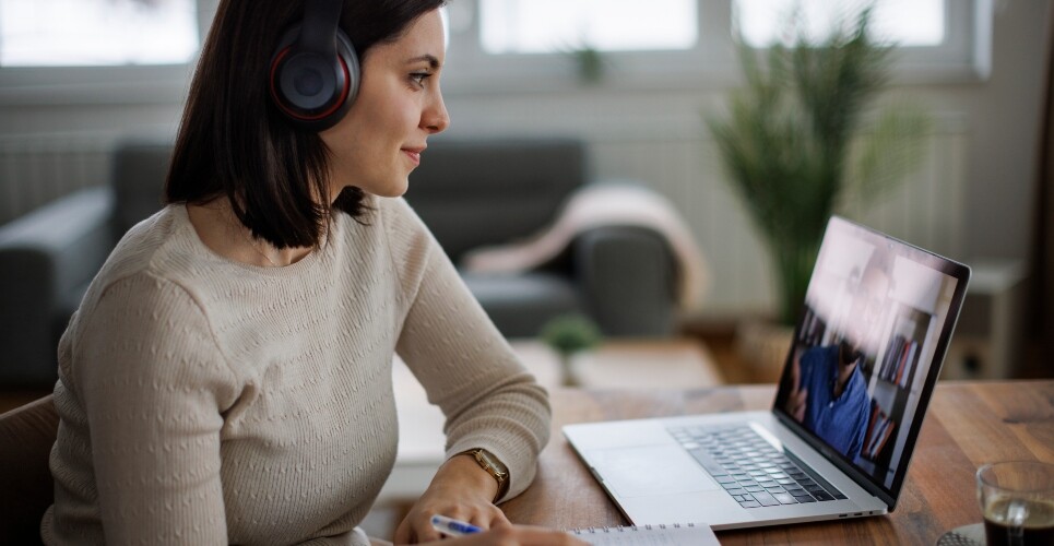 A woman sat at a desk wearing headphones and watching a webinar on her laptop while taking notes in a notebook to illustrate the launch of Hospital Pharmacy Europe's biosimilars virtual study day