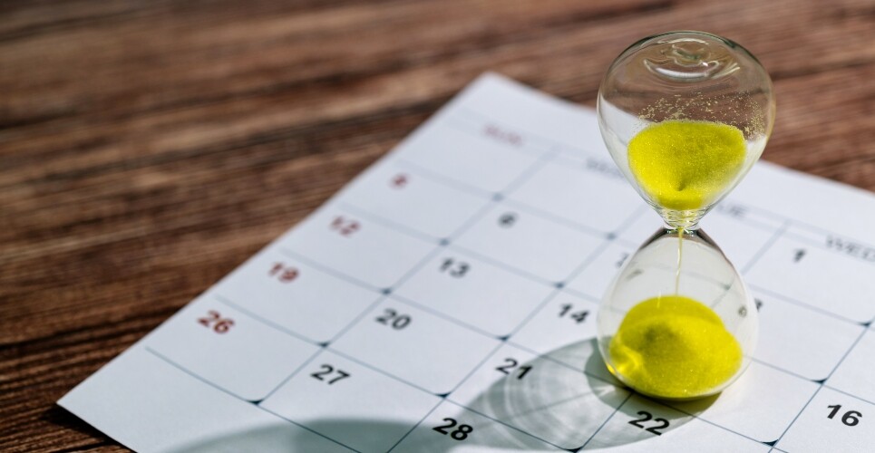 A glass hourglass with yellow sand sitting on a page of a calendar on a wooden table to illustrate the benefits of early biologic initiation in psoriasis
