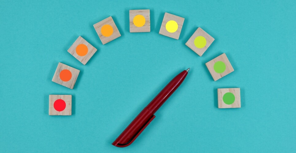 Wooden blocks with coloured dots arranged in a curved traffic-light scale from red to green with a burgundy pen pointing to the yellow to green transition on a turquoise background to illustrate the value of patient-reported outcomes during tildrakizumab psoriasis treatment