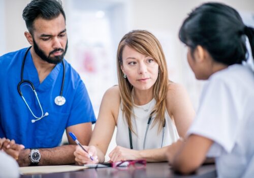 A multi-ethnic group of doctors are indoors in an office. They are gathered around a table for a meeting about ARRS