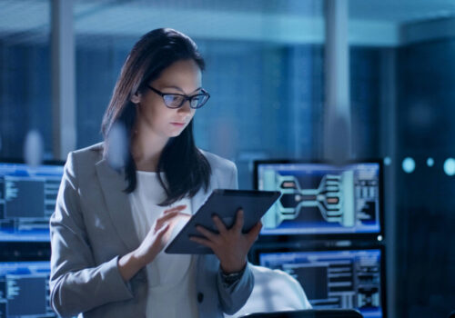 A woman with dark hair and glasses wearing a grey blazer and using an electronic tablet. There are computers in the background.