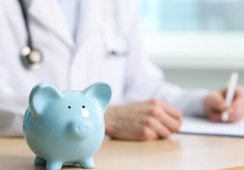 A piggy bank on a table with a doctor writing notes in the background.