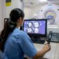 Radiology technician performing brain MRI on patient looking at screen in front of window to next room with patient in scanner as part of neurology investigation