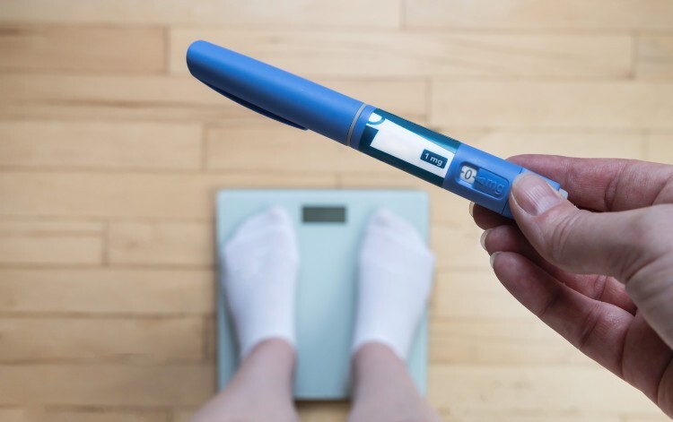 Person standing on weighing scales holding a weight loss injection close to the camera