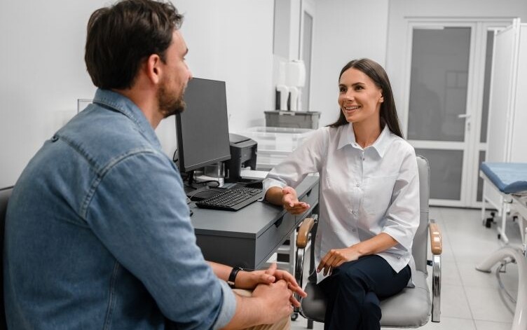 Female healthcare professional in consultation with adult male sitting by desk with computer to depict process of antidepressant medication review by pharmacist