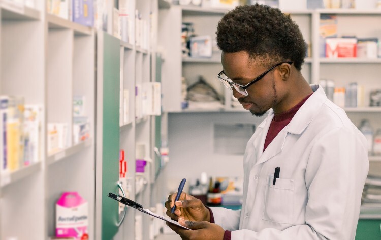 A pharmacist making notes on a clipboard with medicines on the walls in the background.