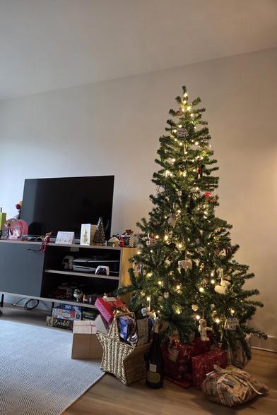 A decorated Christmas tree with lights and ornaments stands next to a TV, with wrapped presents and a basket arranged at its base in a living room.