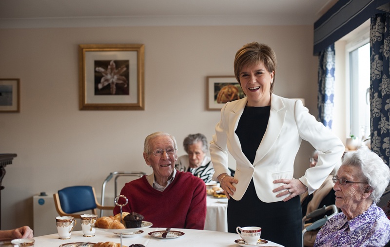 Nicola Sturgeon standing up next to two older people seated at a table with cups of tea and cakes