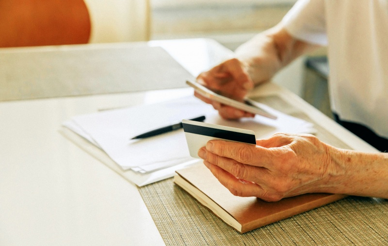 Elderly woman holding a credit card and a phone with some documents laid out on a table in front of her