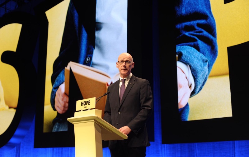 John Swinney speaking at Conference in front of an image of a child reading a book